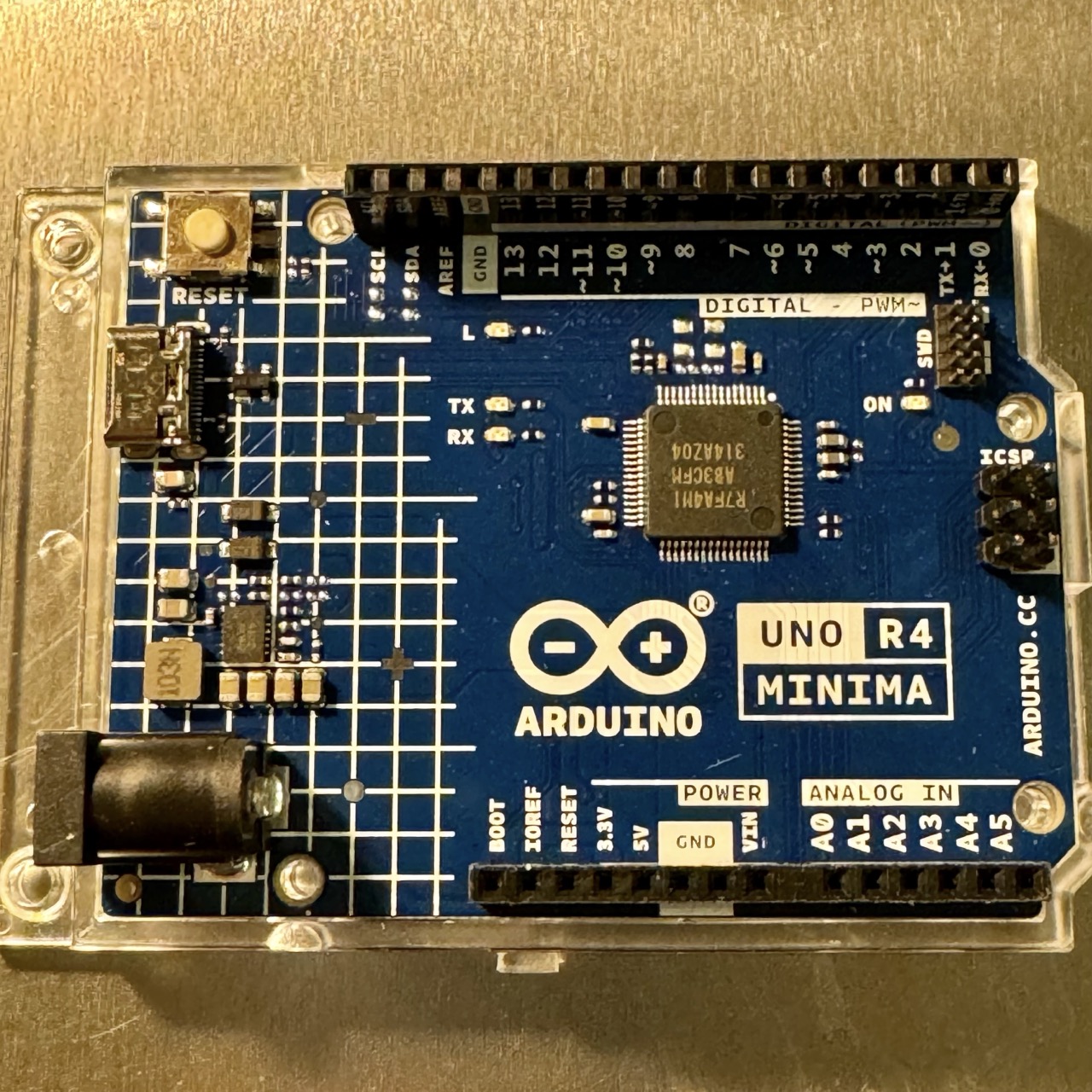 Arduino R4 Minima board with dark-blue PCB, featuring the Renesas RA4M1 microcontroller. The board includes a USB-C connector, multiple digital and analog I/O pins, power supply pins, and the Arduino logo. The layout is compact and designed for both beginners and advanced users in IoT, robotics, and embedded projects.